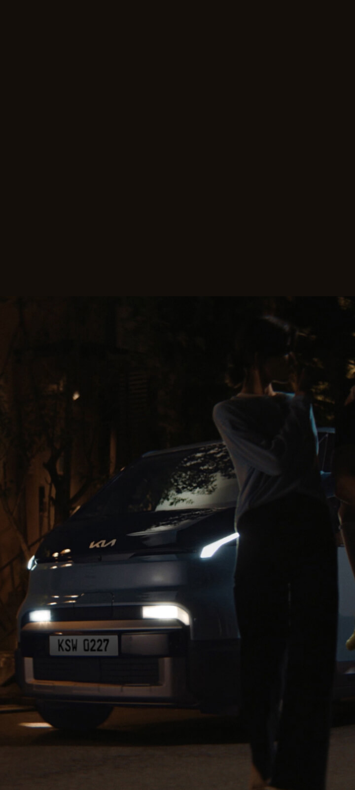 Nächtliche Szene: Der Kia PV5 Passenger mit eingeschaltetem Licht, während eine Familie davor steht.